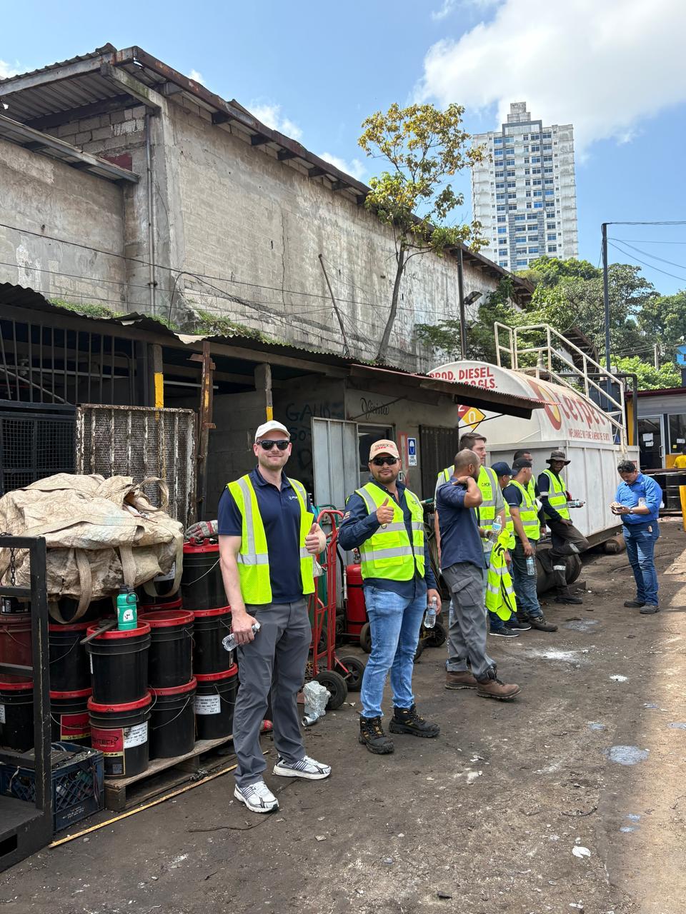 Fuchs, ICP, and yard crew in high-visibility vests standing outside the scrap yard workshop, two of them giving a thumbs up