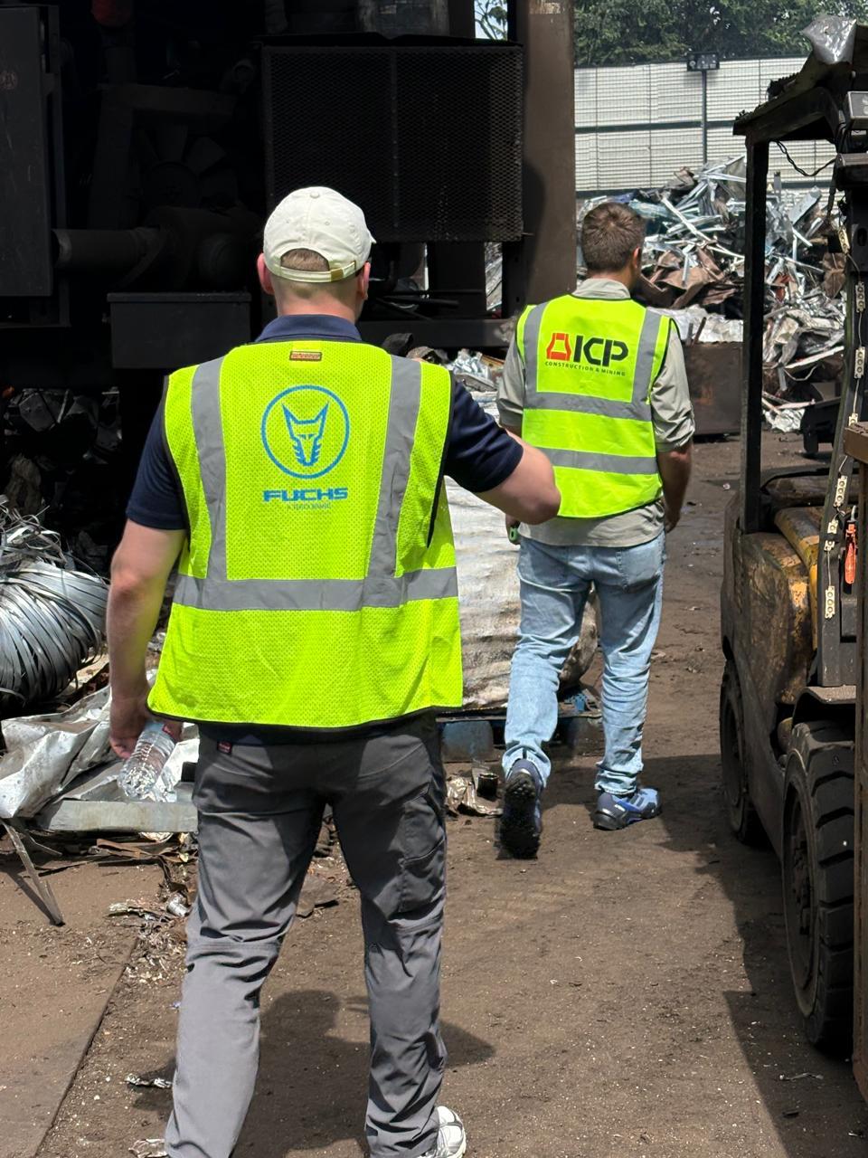 Two men in Fuchs and ICP safety vests walking through the scrap yard past piles of scrap metal and a forklift