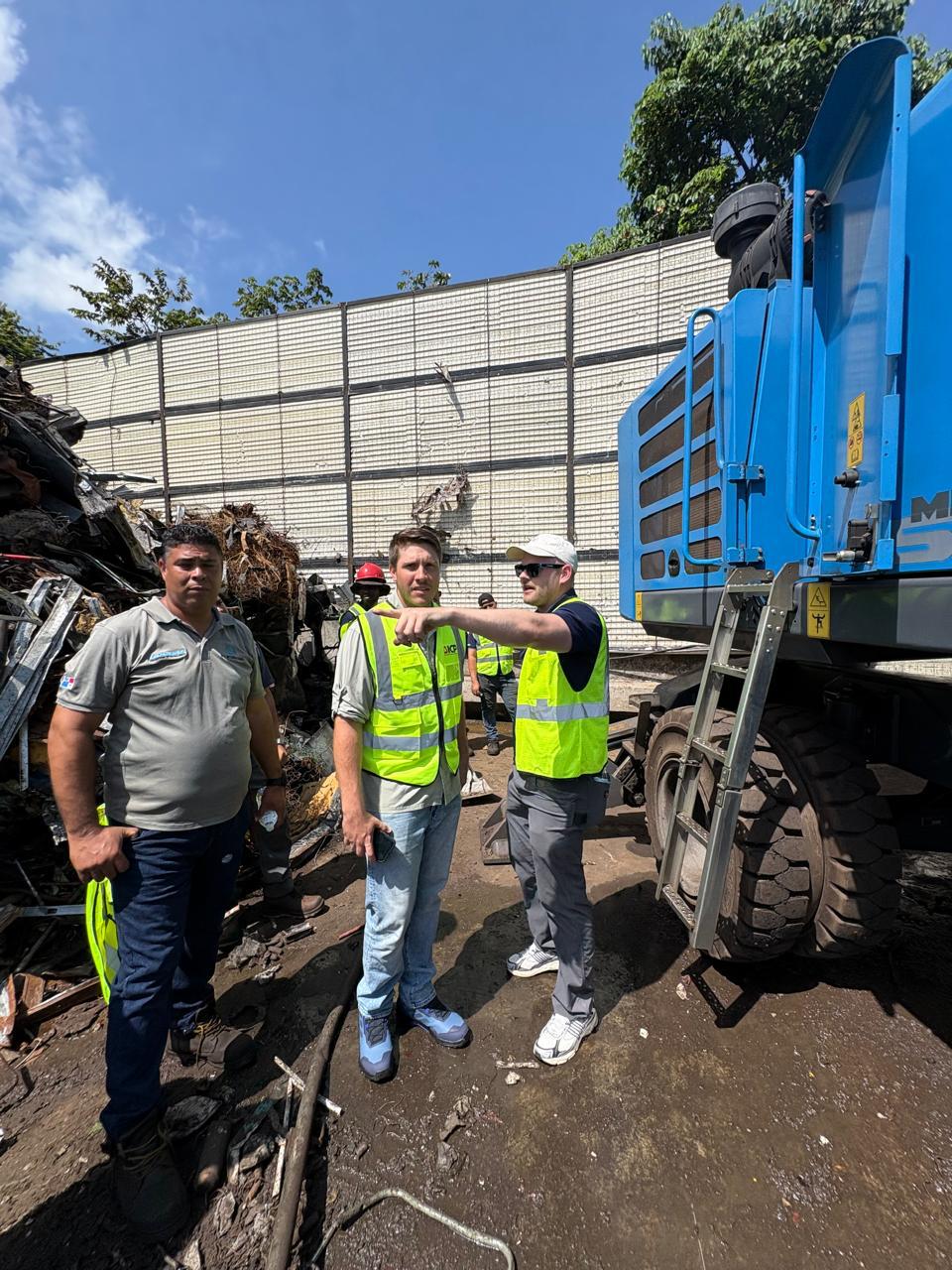 Fuchs representative pointing while explaining the machine to two yard crew members beside a blue Fuchs MHL 350 material handler