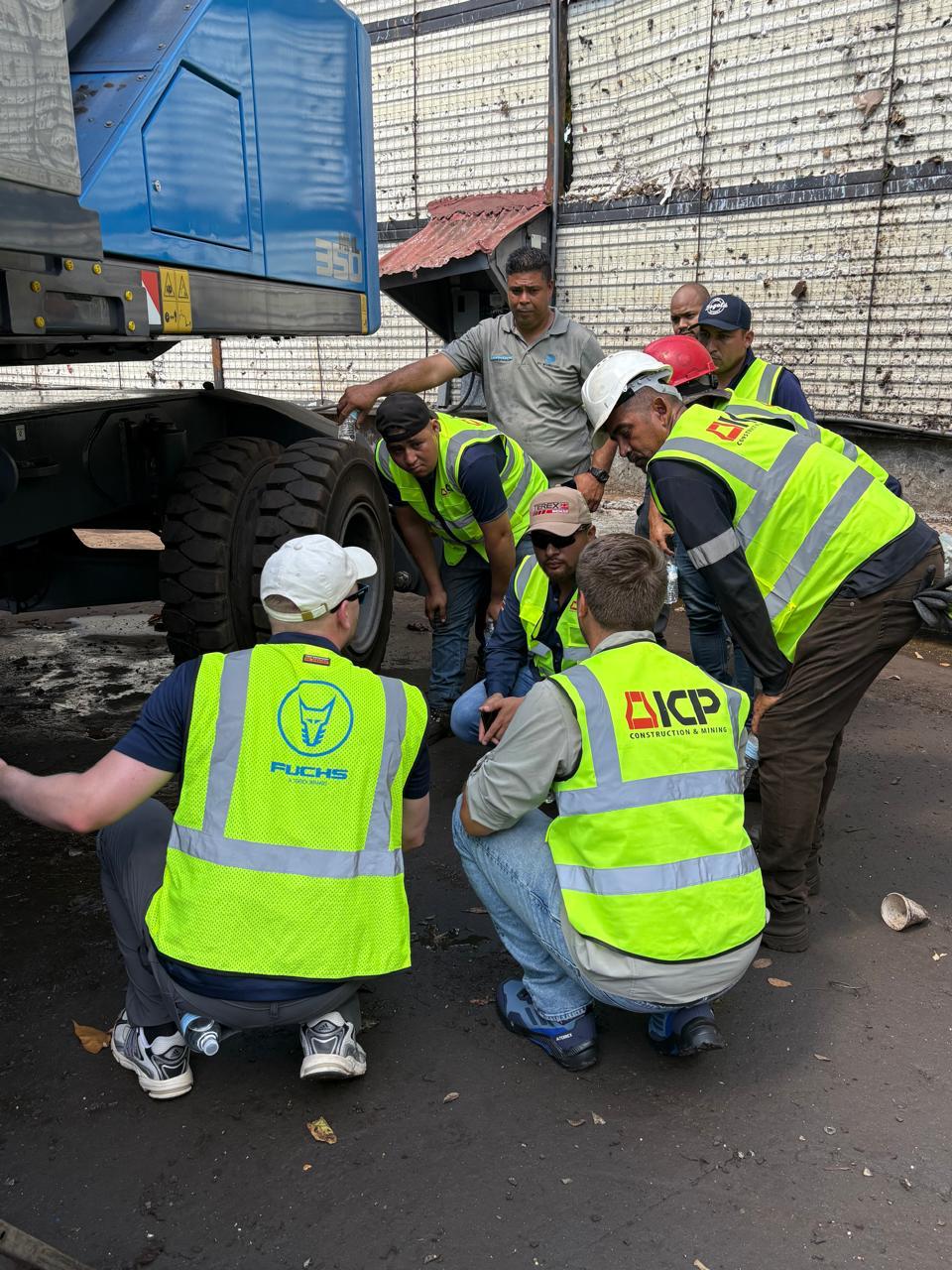 Group of Fuchs, ICP, and yard mechanics in safety vests and hard hats crouched at the wheel and undercarriage of the Fuchs MHL 350