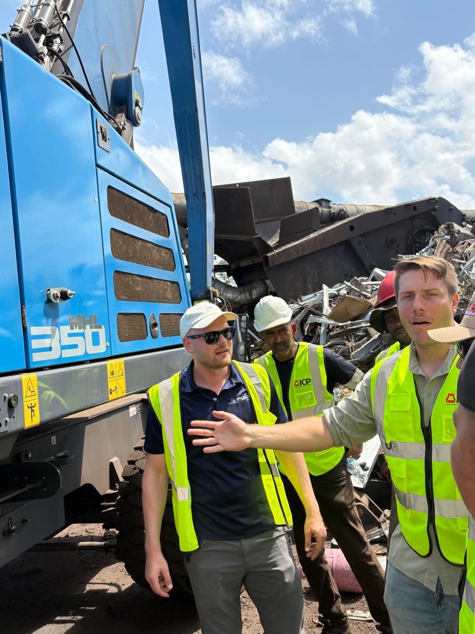 Fuchs and ICP representatives with the yard crew standing in front of the blue Fuchs MHL 350 with a scrap pile behind them