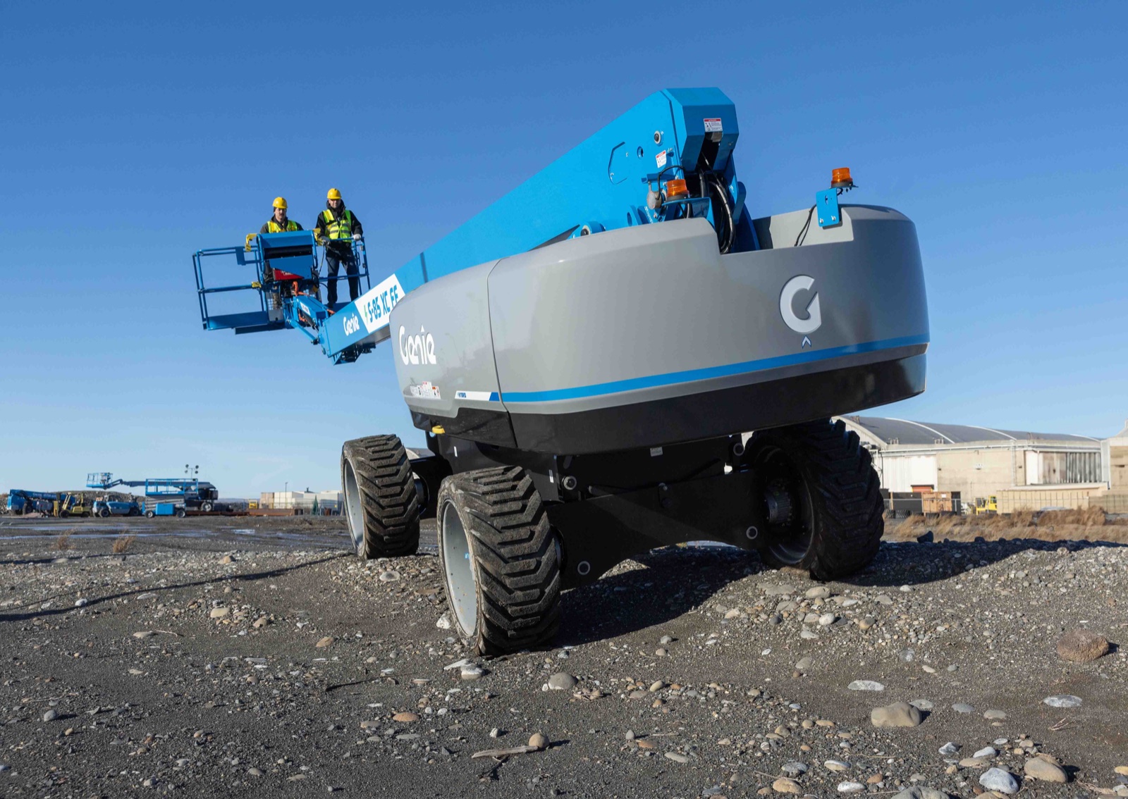 Genie S-85 XC FE hybrid boom lift driving over rough gravel terrain with two workers in the platform
